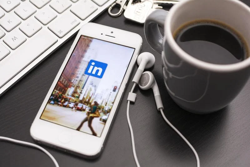 An iPhone loading the LinkedIN app with a cup of coffee resting on a wooden table.