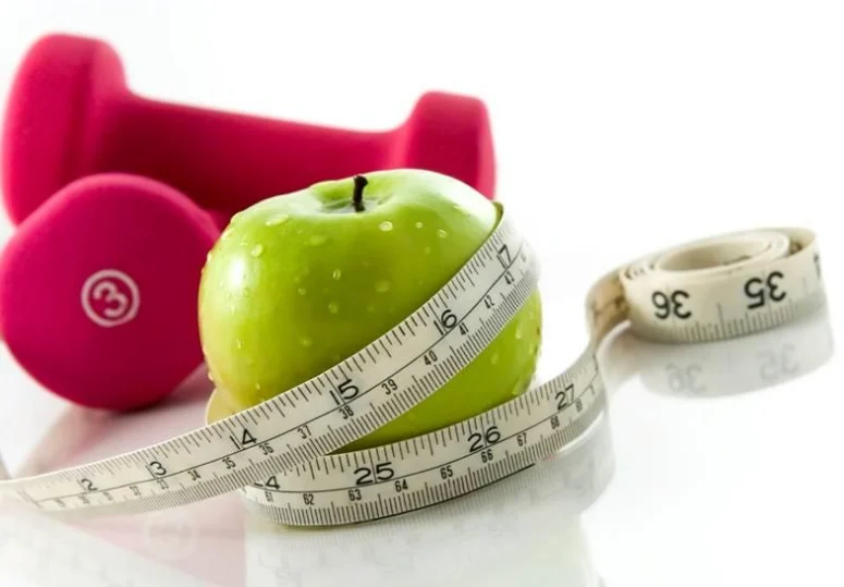 An apple with a measuring tape wrapped around it and red dumbells in the background.
