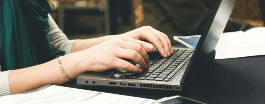 A person sitting on a chair with their laptop resting on the table as they type.