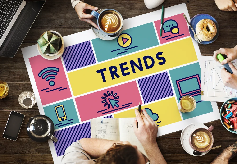 Colourful piece of paper with the word "Trends" written on top with people surrounding it with cups of coffee