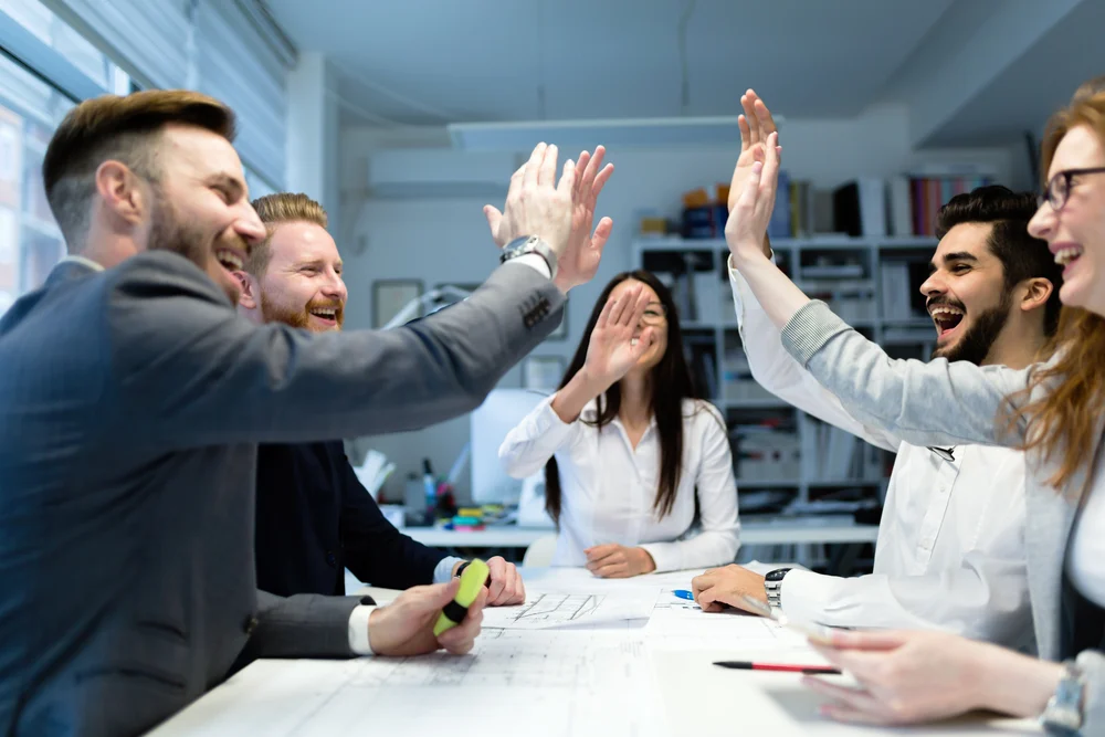 Photograph of business people surrounding a white table high fiving due to winning business contracts