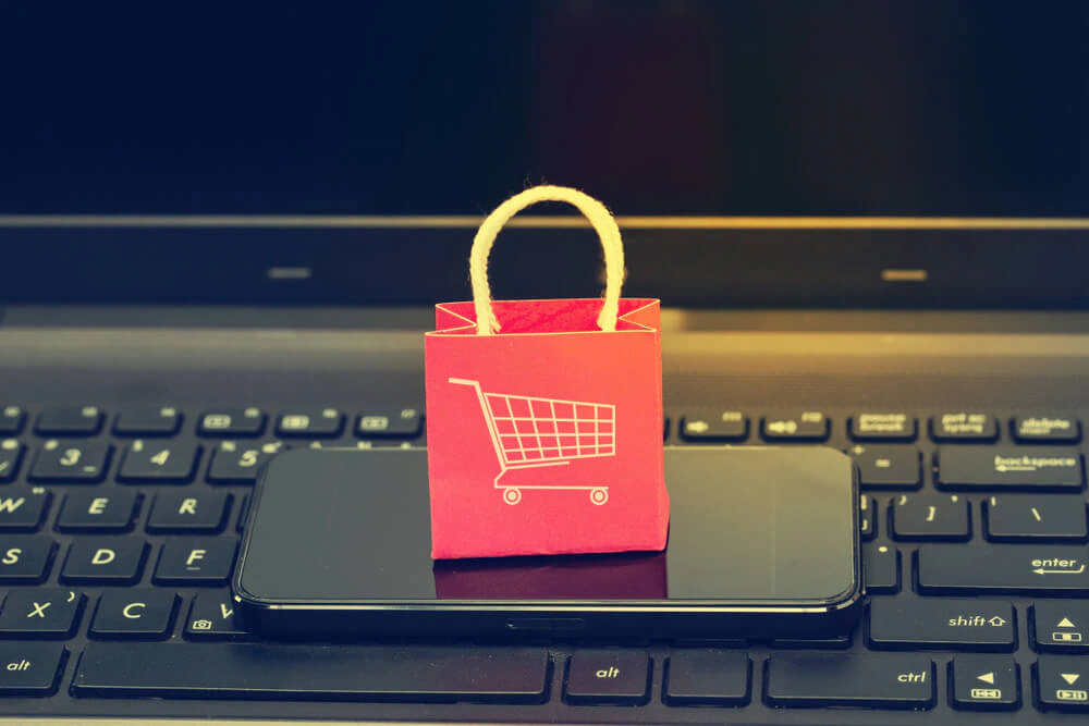 Miniature red shopping bag with a trolley icon resting on a smartphone and laptop keyboard