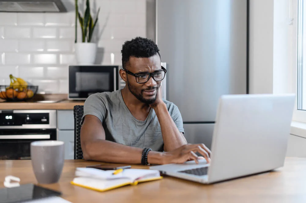 A tired male employee working from home on a laptop sat at his kitchen table looking stressed 