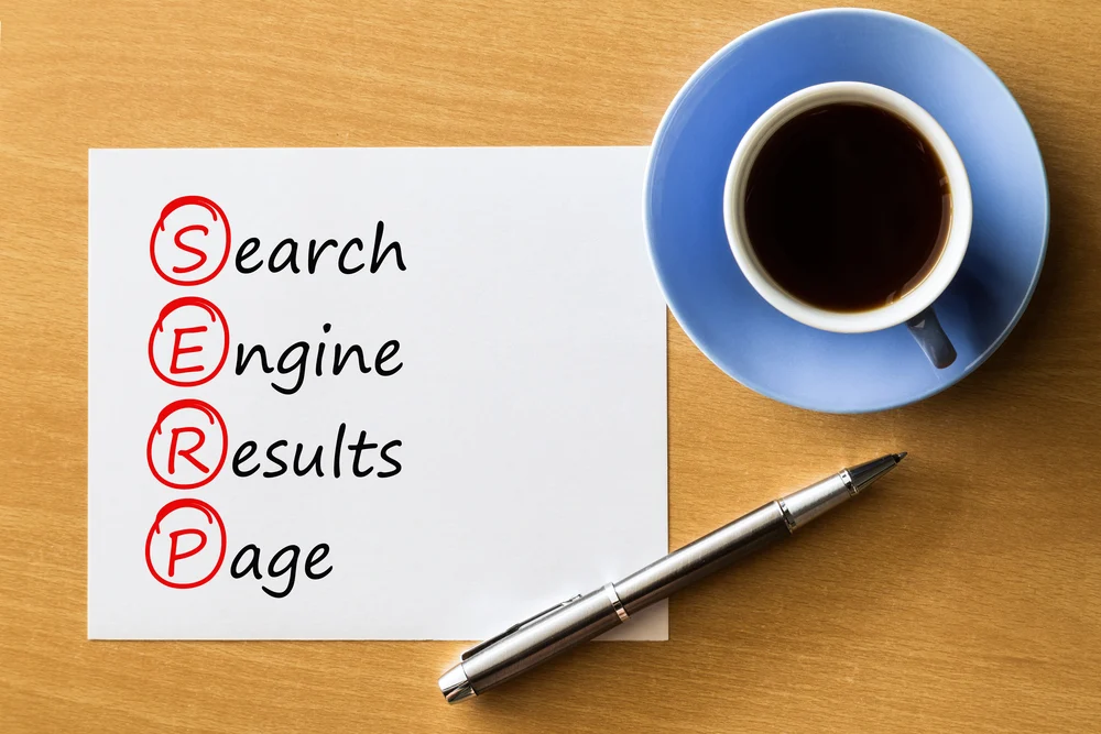A wooden table with a cup of coffee and a saucer next to a piece of paper showing that SERP stands for; serch engine results page.