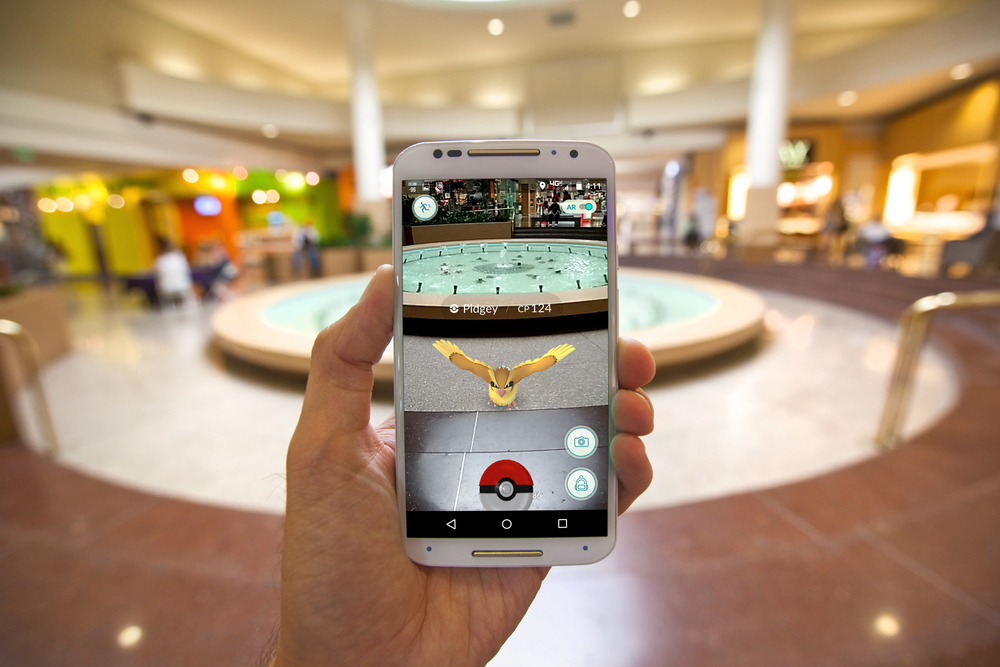A hand holding a phone displaying the Pokémon GO game, with a blurred city square in the background.