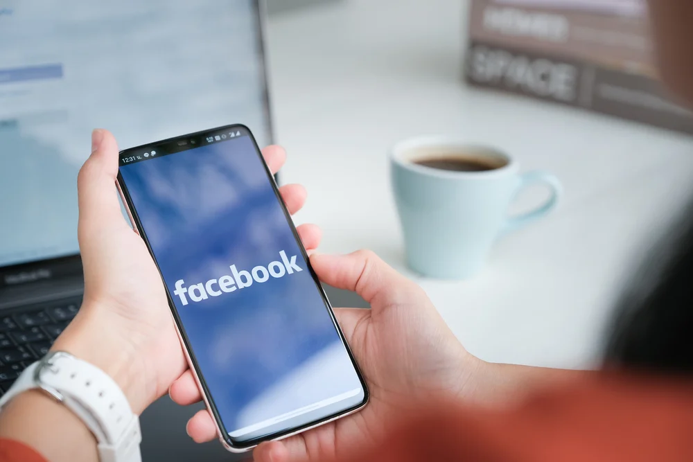 The Facebook app being opened on an iPhone with a social media assistant preparing to schedule a post.