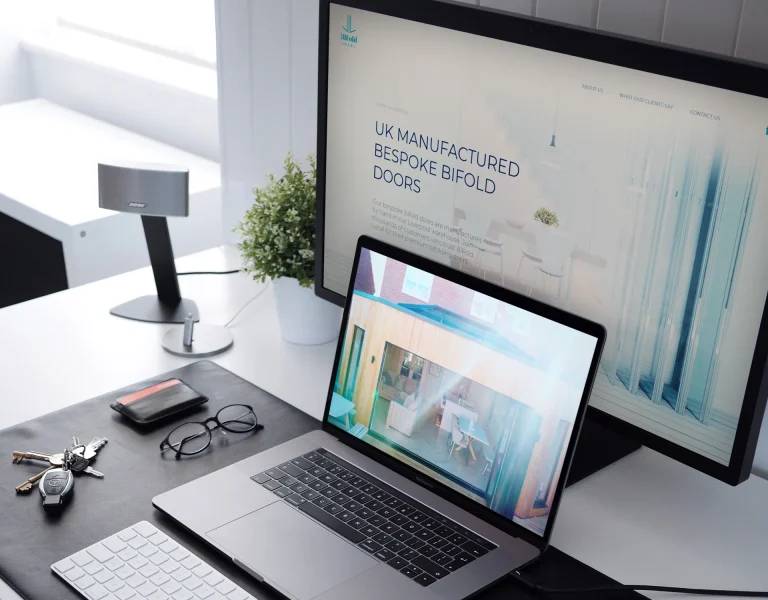 Image of a desk with a desktop computer showing the BiFold Local website and a laptop in front showing an image of BiFold locals bifold doors