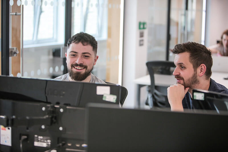Two Castle Int. colleagues chatting behind their monitors in the Liverpool office