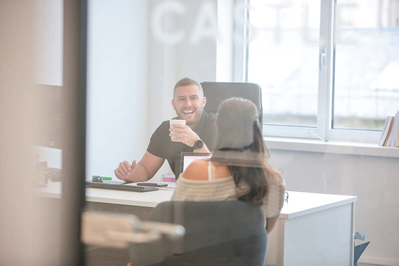 Two Castle Int. colleagues sharing a laugh over coffee at a desk in the Liverpool office