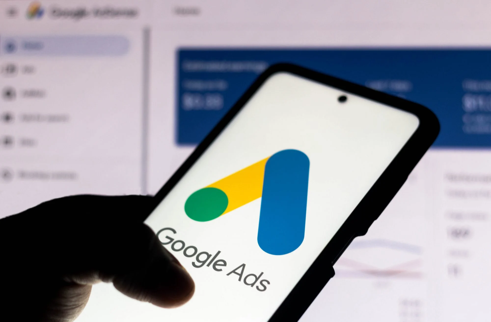 Hand holding a smartphone showing the Google Ads logo in front of a campaign dashboard