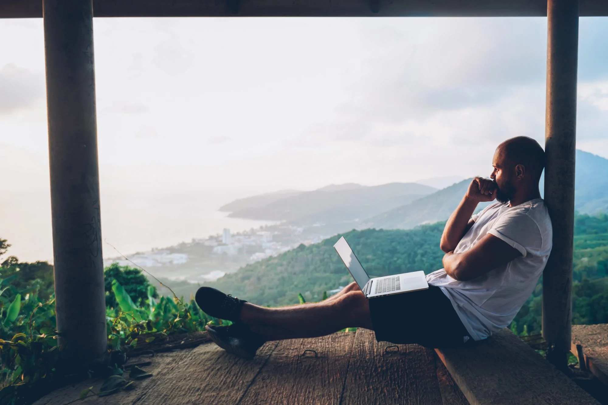 Young male travel blogger guy catch inspiration writing article on laptop during vacation holidays in beautiful location, looking out thoughtfully onto the natural landscape