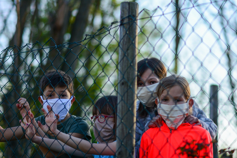 Children wearing face masks looking through a chain-link fence during the pandemic