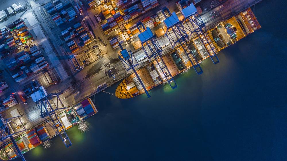 Aerial top view of a container cargo ship working at night. It's stopped at the docks next to row after row of shipping containers. 