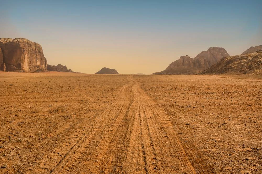 An arid landscape of red sand stretches for miles, with a backdrop of mountains and blue sky. 