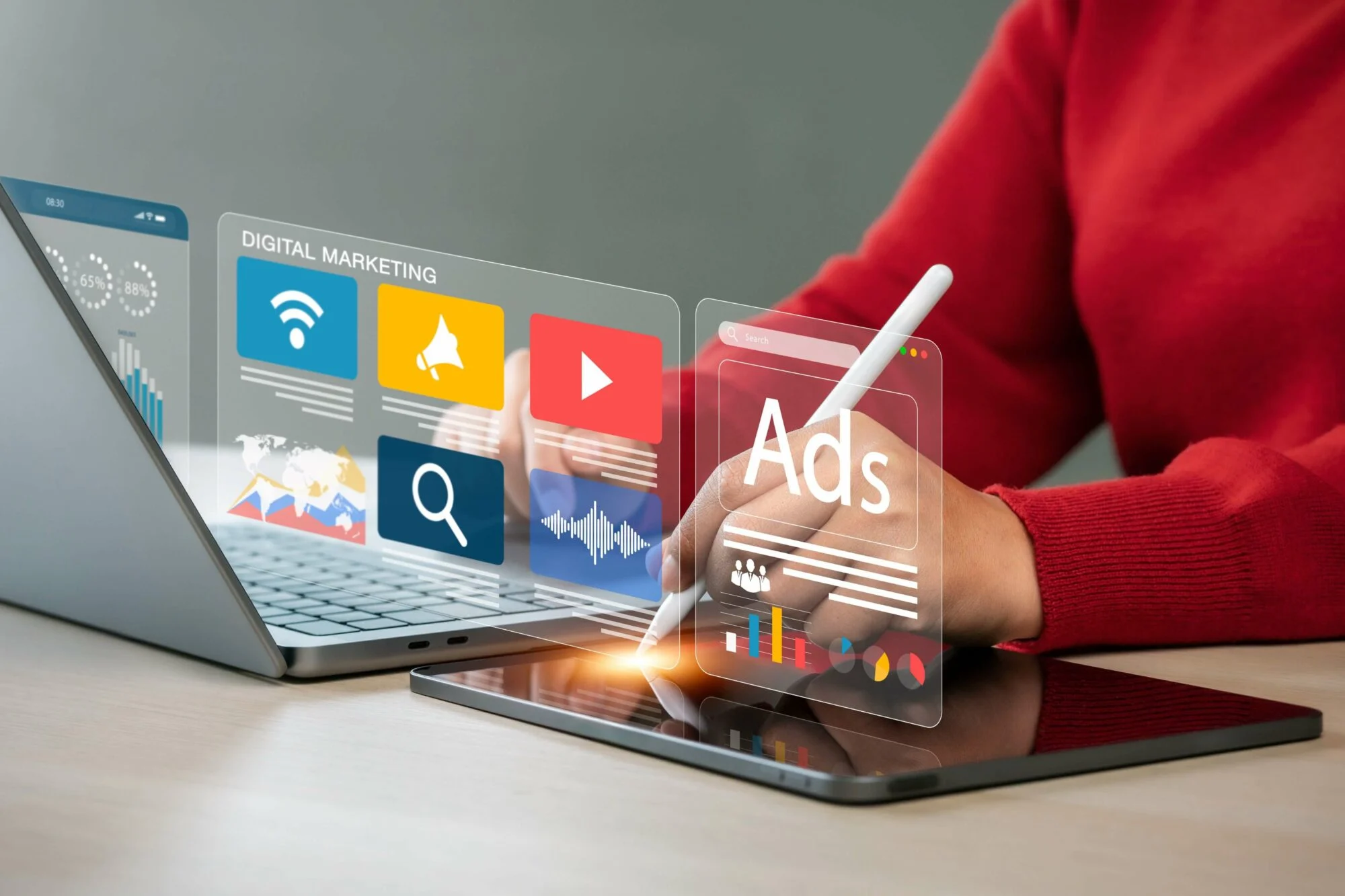 An image of a person in a red jumper sat behind a desk with a laptop and tablet in front of them. They're typing on the tablet with a white stylus and hovering above the laptop and tablet are graphics symbolising ads and digital marketing.