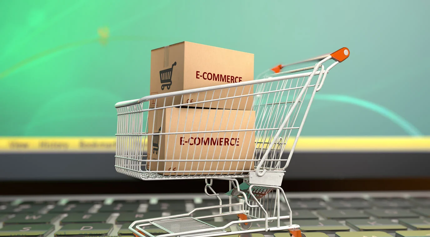 An image of a shopping trolley on top of a laptop keyboard with two brown boxes inside it with the text 'E-COMMERCE' written on the two brown boxes. 