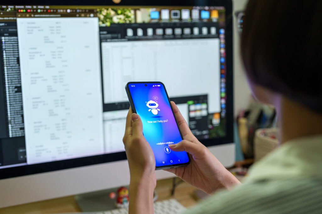 User interacting with an AI chatbot and voice assistant on a smartphone in front of a computer workstation, demonstrating artificial intelligence technology in a work environment