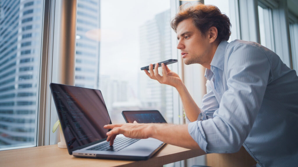 Professional using a smartphone voice assistant alongside a laptop in a modern office, showcasing AI-powered voice commands and workplace productivity technology.