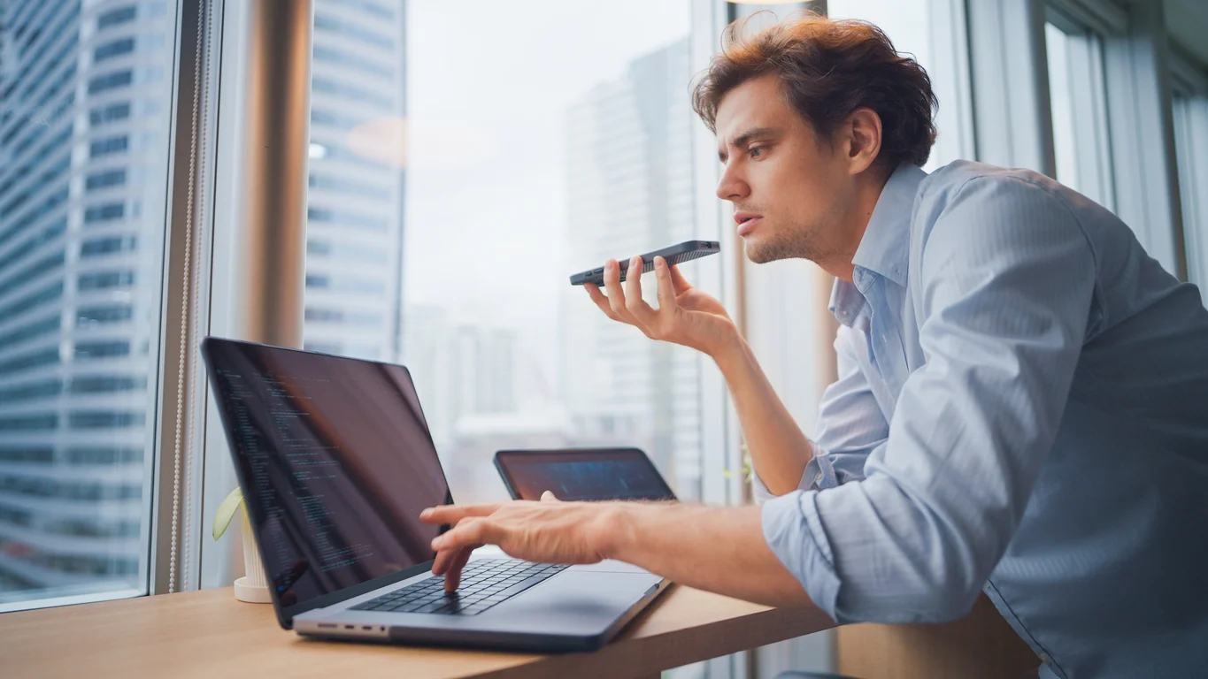 Professional using a smartphone voice assistant alongside a laptop in a modern office, showcasing AI-powered voice commands and workplace productivity technology.